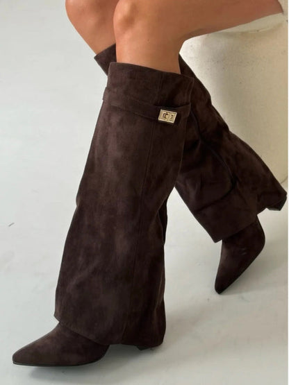 Stivali alti in pelle scamosciata marrone con tacco basso e fibbia dorata, indossati su gambe incrociate. Moda autunnale, calzature eleganti.
