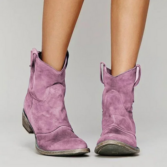 Stivali texani viola in pelle scamosciata, tacco basso, indossati su sfondo grigio. Scarpe moda donna, stile western, calzature trendy.