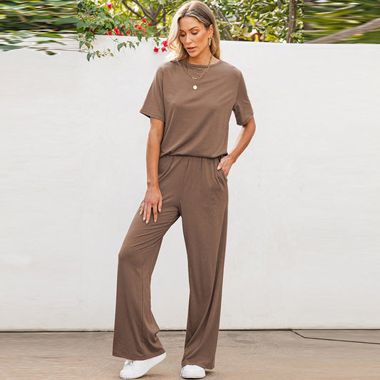 Donna in tuta marrone casual, con pantaloni larghi e maglietta a maniche corte, indossa scarpe bianche. Sfondo con piante verdi e muro bianco.