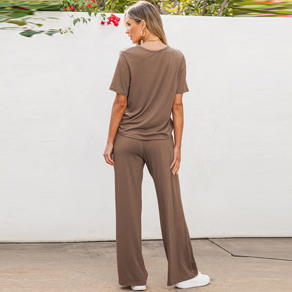 Donna di spalle in tuta marrone, composta da maglietta e pantaloni larghi, in piedi su pavimento esterno. Sfondo bianco con piante verdi e fiori rossi.