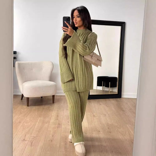 Donna in abbigliamento invernale verde, posa davanti a uno specchio. Indossa un maglione e pantaloni a coste, con borsa beige. Stanza moderna.