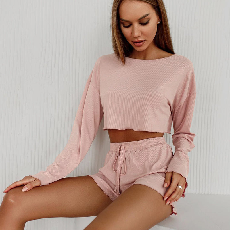 Donna in abbigliamento lounge rosa, top a maniche lunghe e shorts, seduta su sfondo bianco. Moda casual, stile confortevole, tendenze 2023.