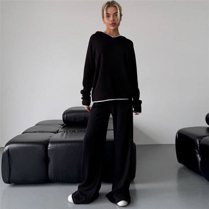 Donna in abbigliamento casual nero, felpa e pantaloni larghi, posa in un salotto moderno con divano in pelle nera. Stile minimalista, moda urbana.