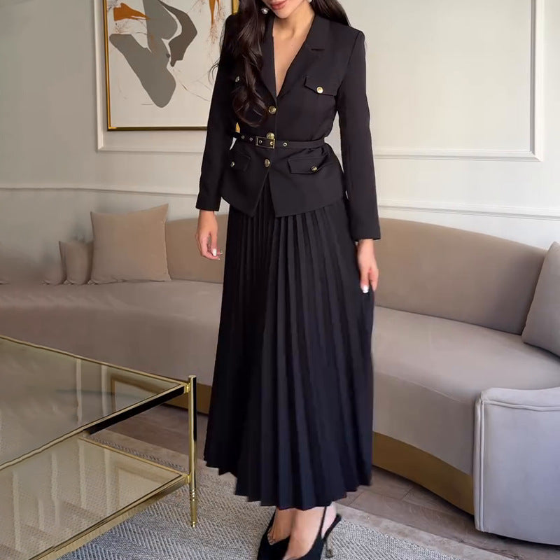 Donna in elegante abito nero con giacca e gonna plissettata, in un salotto moderno con divano beige e tavolino in vetro. Moda femminile chic.