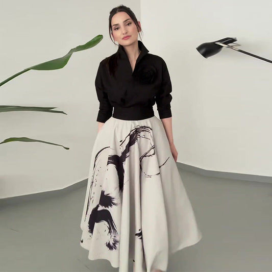 Donna con camicia nera e gonna bianca con stampa artistica, in un ambiente minimalista con pianta e lampada moderna. Moda elegante e contemporanea.