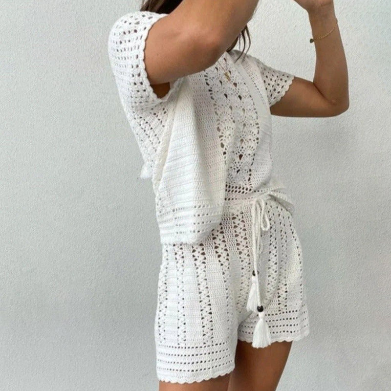 Donna in abito estivo bianco all'uncinetto, con top e pantaloncini coordinati, posa contro un muro bianco. Moda estiva, abbigliamento casual.