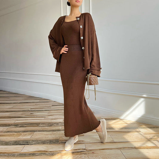 Donna in abito lungo marrone con cardigan abbinato, borsa a tracolla trapuntata e scarpe beige su pavimento in legno. Moda autunnale elegante.