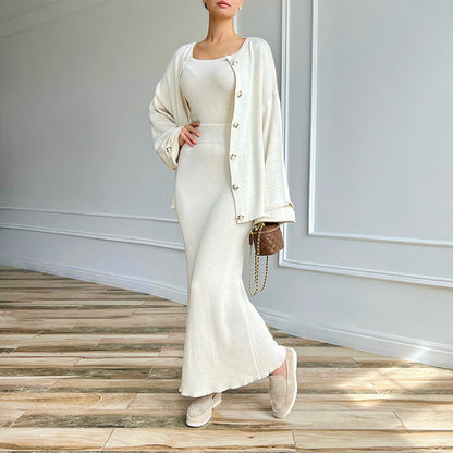 Donna in abito lungo bianco elegante, cardigan abbinato, borsa marrone, scarpe beige, posa su pavimento in legno. Moda femminile chic e sofisticata.