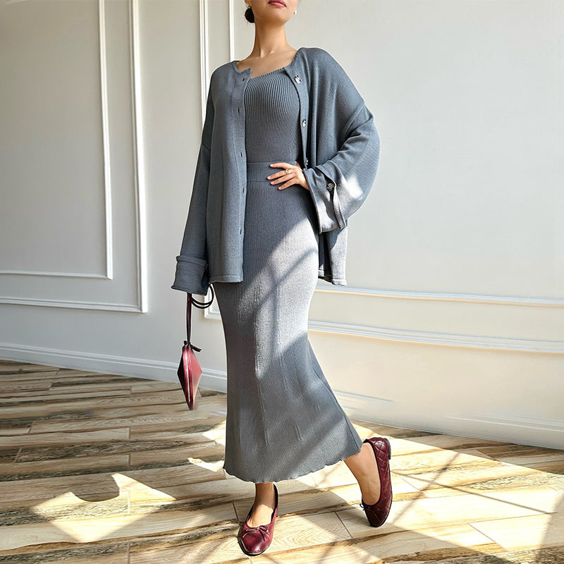Donna in abito grigio elegante con cardigan abbinato, borsa rossa e mocassini, posa su pavimento in legno. Moda autunnale chic e sofisticata.