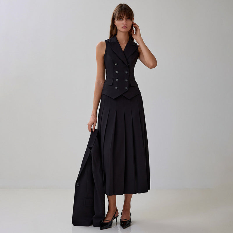 Donna in abito nero elegante con gilet e gonna lunga a pieghe, tacchi alti, su sfondo neutro. Moda femminile, abbigliamento formale, stile sofisticato.