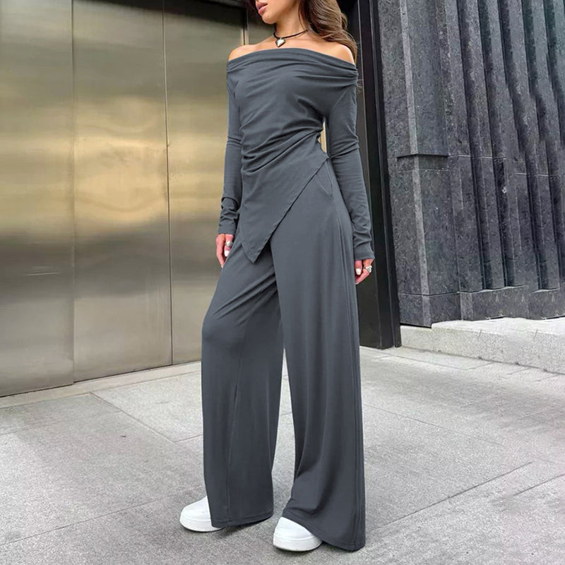 Donna con tuta grigia elegante, spalle scoperte, pantaloni larghi, scarpe bianche, sfondo urbano moderno. Moda femminile contemporanea.