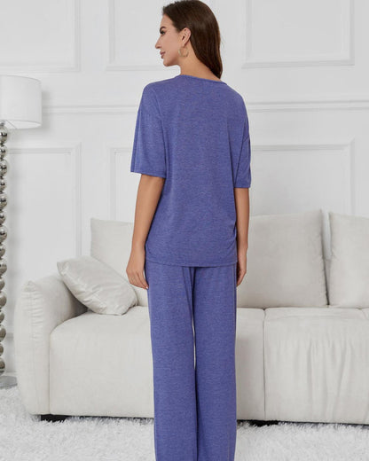 Donna in pigiama viola, vista posteriore, in salotto moderno con divano bianco e lampada. Abbigliamento comodo per casa, moda donna casual.