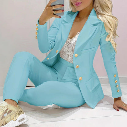 Donna in elegante completo azzurro con bottoni dorati, seduta su pavimento chiaro. Indossa scarpe da ginnastica beige e top in pizzo bianco.