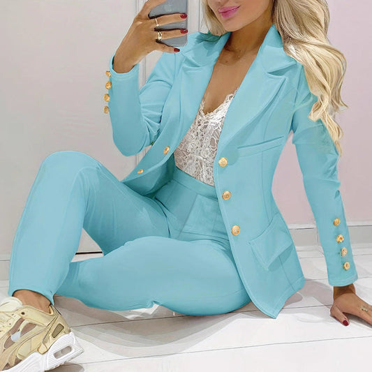 Donna in elegante completo azzurro con bottoni dorati, seduta su pavimento chiaro. Indossa scarpe da ginnastica beige e top in pizzo bianco.
