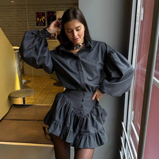 Donna in abito grigio scuro con maniche a sbuffo e gonna a balze, posa vicino a una finestra. Moda autunnale elegante e moderna.