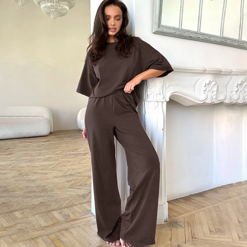Donna in salotto elegante indossa tuta marrone comoda, appoggiata a camino bianco. Moda casual, arredamento moderno, pavimento in parquet.