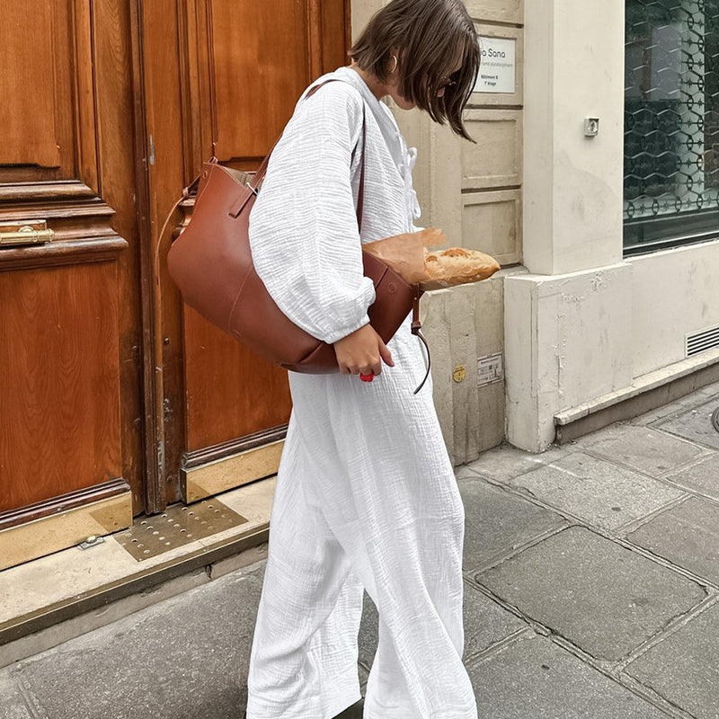 Donna in abito bianco porta borsa marrone con baguette, cammina su marciapiede cittadino. Moda urbana, stile casual, shopping.