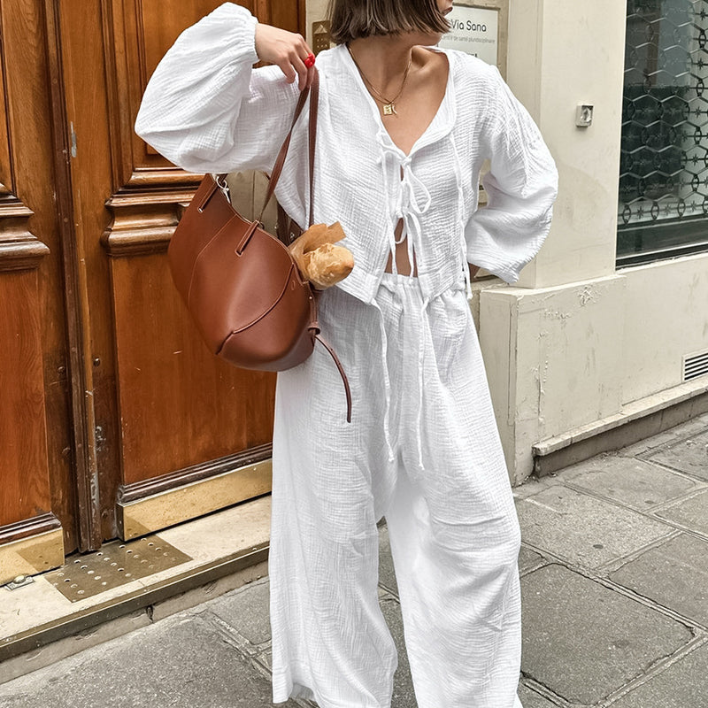 Donna con abito bianco estivo, borsa marrone e baguette, cammina su marciapiede cittadino. Moda casual, street style, outfit estivo elegante.