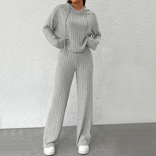 Donna in tuta grigia a coste, con cappuccio e pantaloni larghi, su sfondo bianco. Moda casual, abbigliamento comodo, stile autunnale.
