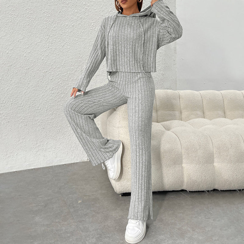 Donna in tuta grigia a coste, posa su divano bianco. Abbigliamento casual, moda autunnale, comfort, stile moderno. Scarpe da ginnastica bianche.