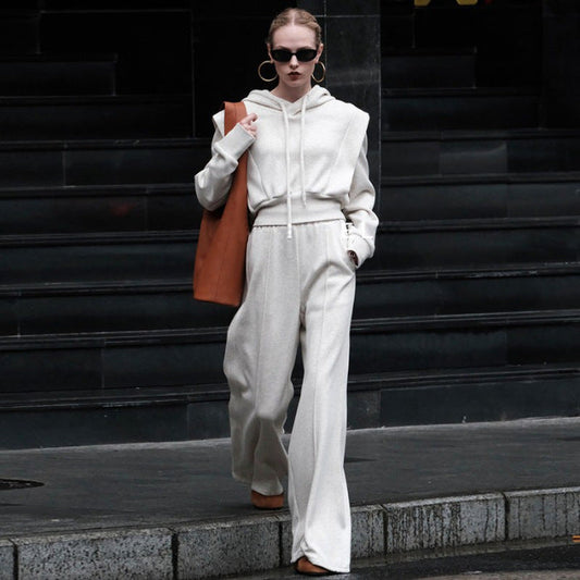 Donna con occhiali da sole indossa tuta bianca elegante e borsa marrone, cammina su marciapiede urbano. Moda autunnale, stile casual chic.