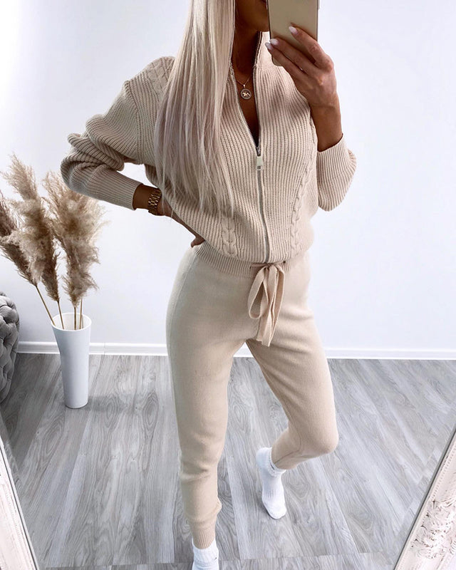 Donna in tuta beige a maglia, con zip e cintura, scatta selfie in stanza moderna con pavimento in legno e piante decorative. Moda autunnale casual.