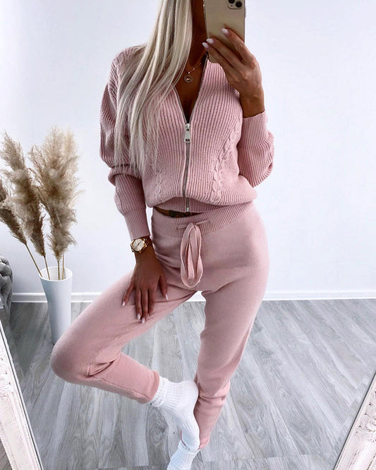 Donna in tuta rosa con zip, posa davanti a uno specchio. Indossa calzini bianchi, orologio dorato. Sfondo con piante decorative. Moda casual elegante.