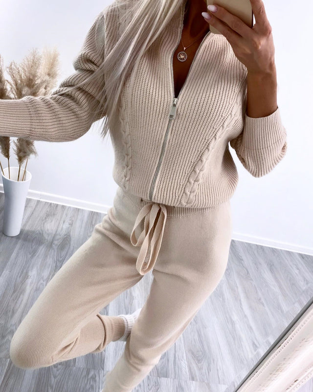 Donna in tuta beige a maglia con zip, posa davanti a uno specchio. Moda autunnale, abbigliamento casual elegante, interni moderni.