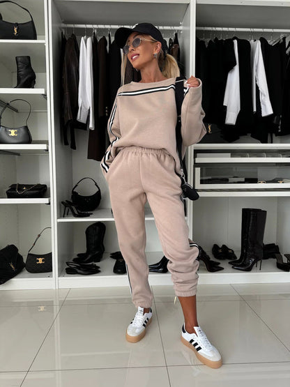Donna in tuta beige con strisce nere, cappellino nero e occhiali da sole, posa in un armadio moderno con scarpe e abbigliamento elegante.