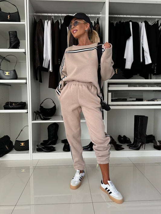 Donna in tuta beige con strisce nere, cappellino nero e occhiali da sole, posa in un armadio moderno con scarpe e abbigliamento elegante.
