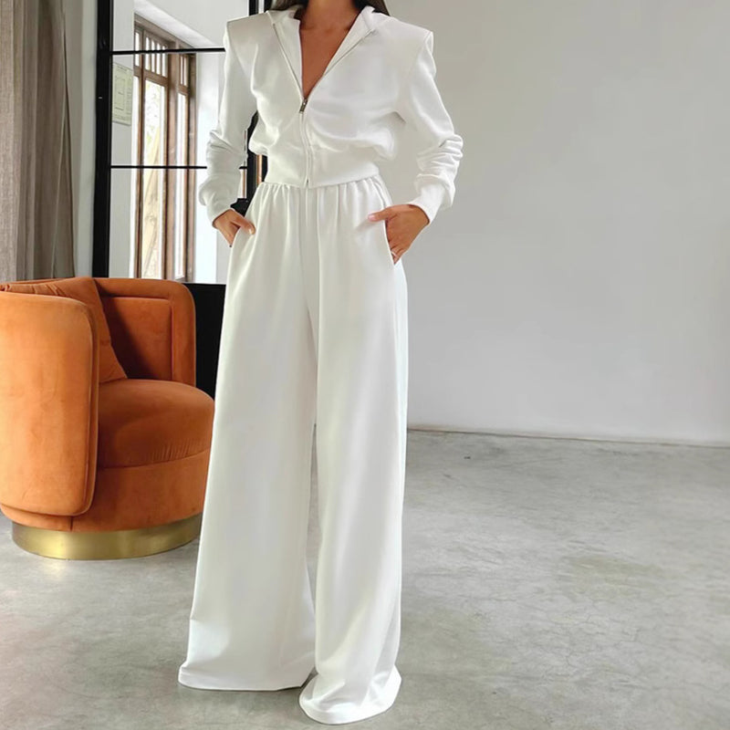 Tuta bianca elegante con zip, maniche lunghe e pantaloni larghi, indossata in un salotto moderno con poltrona arancione. Moda donna contemporanea.