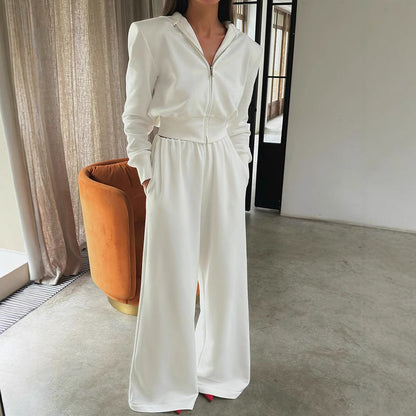 Donna in tuta bianca elegante, con cappuccio e pantaloni larghi, in un salotto moderno con poltrona arancione e pavimento in cemento.