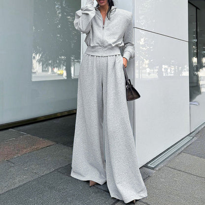 Donna in tuta grigia oversize, con pantaloni larghi e giacca con zip, tiene una borsa nera. Look casual chic, moda urbana autunnale.