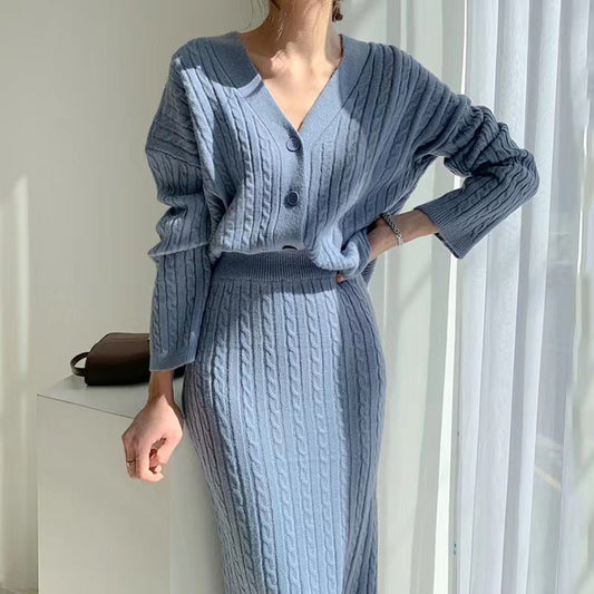 Donna con abito in maglia blu, cardigan a coste e gonna coordinata, posa vicino a una finestra luminosa. Moda autunnale elegante e confortevole.