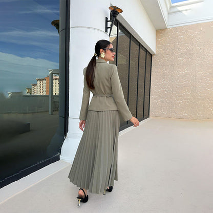 Donna elegante in abito verde oliva con giacca e gonna plissettata, occhiali da sole, cammina su terrazza moderna. Moda femminile, stile sofisticato.