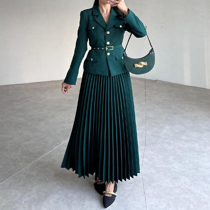 Donna in abito verde elegante con giacca a bottoni e gonna plissettata, borsa a tracolla nera, scarpe a punta. Moda autunnale chic.