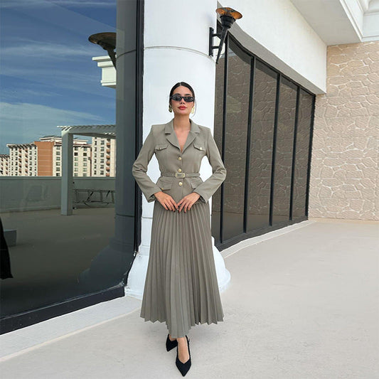 Donna elegante in abito lungo beige con giacca e gonna plissettata, posa su terrazza moderna. Moda femminile, stile sofisticato, abbigliamento chic.