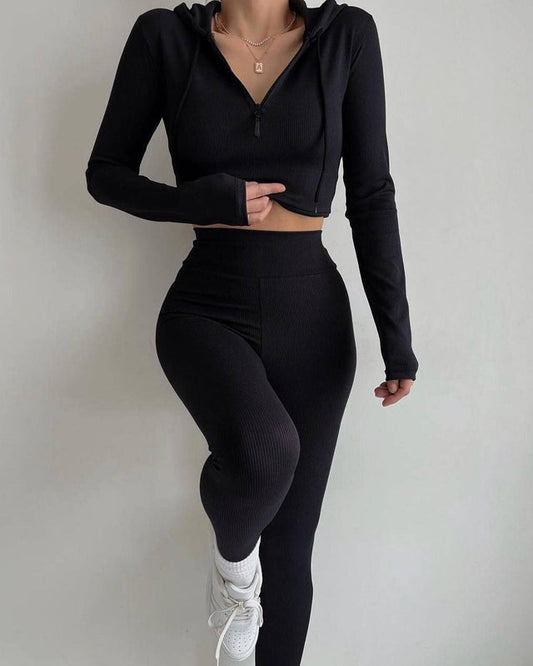 Donna in abbigliamento sportivo nero, con top a maniche lunghe e leggings, posa su sfondo bianco. Moda fitness, abbigliamento activewear elegante.
