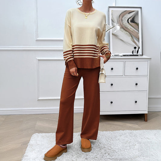 Donna in salotto moderno indossa maglione beige a righe e pantaloni marroni, con scarpe beige. Accanto, cassettiera bianca e quadro astratto.