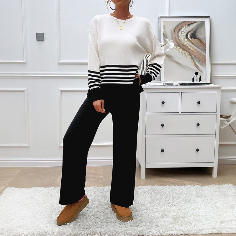 Donna in maglione bianco e nero a righe, pantaloni neri, su tappeto bianco in soggiorno moderno. Arredamento minimalista, cassettiera e quadro astratto.