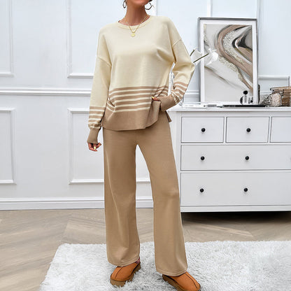 Donna in salotto indossa completo beige a righe, maglione e pantaloni larghi, su tappeto bianco. Arredamento moderno con cassettiera e decorazioni.