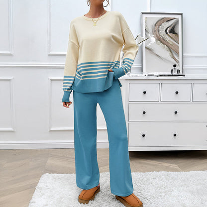Donna con maglione beige e blu a righe, pantaloni blu, scarpe marroni, in una stanza moderna con cassettiera bianca e quadro astratto.