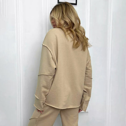 Donna di spalle con capelli biondi indossa tuta beige, composta da felpa e pantaloni, davanti a parete bianca. Moda casual, abbigliamento comodo.
