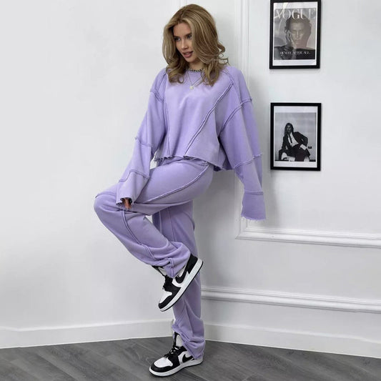 Donna in tuta sportiva lilla, posa in una stanza moderna con riviste Vogue sullo sfondo. Indossa sneakers Nike bianche e nere. Moda casual elegante.