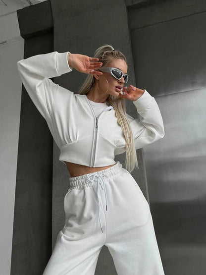 Donna in abbigliamento sportivo bianco, posa moderna, occhiali da sole, capelli lunghi biondi, sfondo urbano grigio. Moda streetwear femminile.