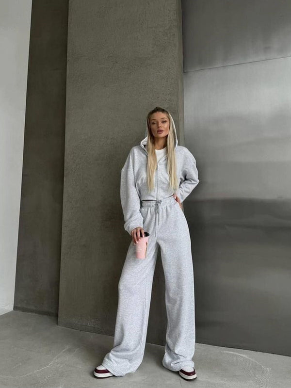 Giovane donna in tuta grigia con cappuccio, pantaloni larghi e scarpe da ginnastica, tiene una bottiglia rosa. Sfondo moderno e minimalista.