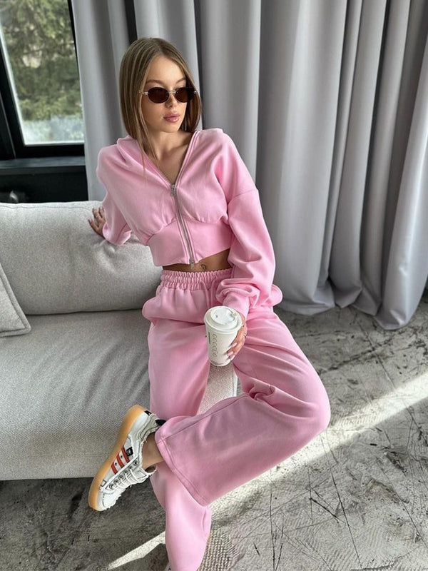 Donna con occhiali da sole indossa tuta rosa e tiene caffè, seduta su divano moderno. Moda casual, abbigliamento comodo, stile urbano.