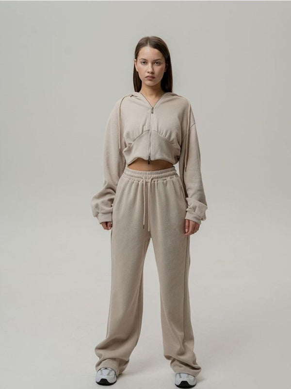 Giovane donna in tuta sportiva beige, felpa con cappuccio e pantaloni larghi, posa su sfondo neutro. Abbigliamento casual, moda donna, comfort.