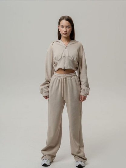 Giovane donna in tuta sportiva beige, felpa con cappuccio e pantaloni larghi, posa su sfondo neutro. Abbigliamento casual, moda donna, comfort.