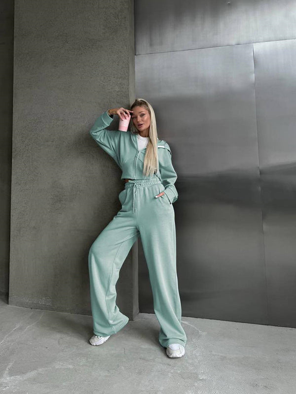 Donna in tuta sportiva verde menta, appoggiata a un muro grigio, con bicchiere rosa in mano. Moda casual, abbigliamento comodo, stile urbano.